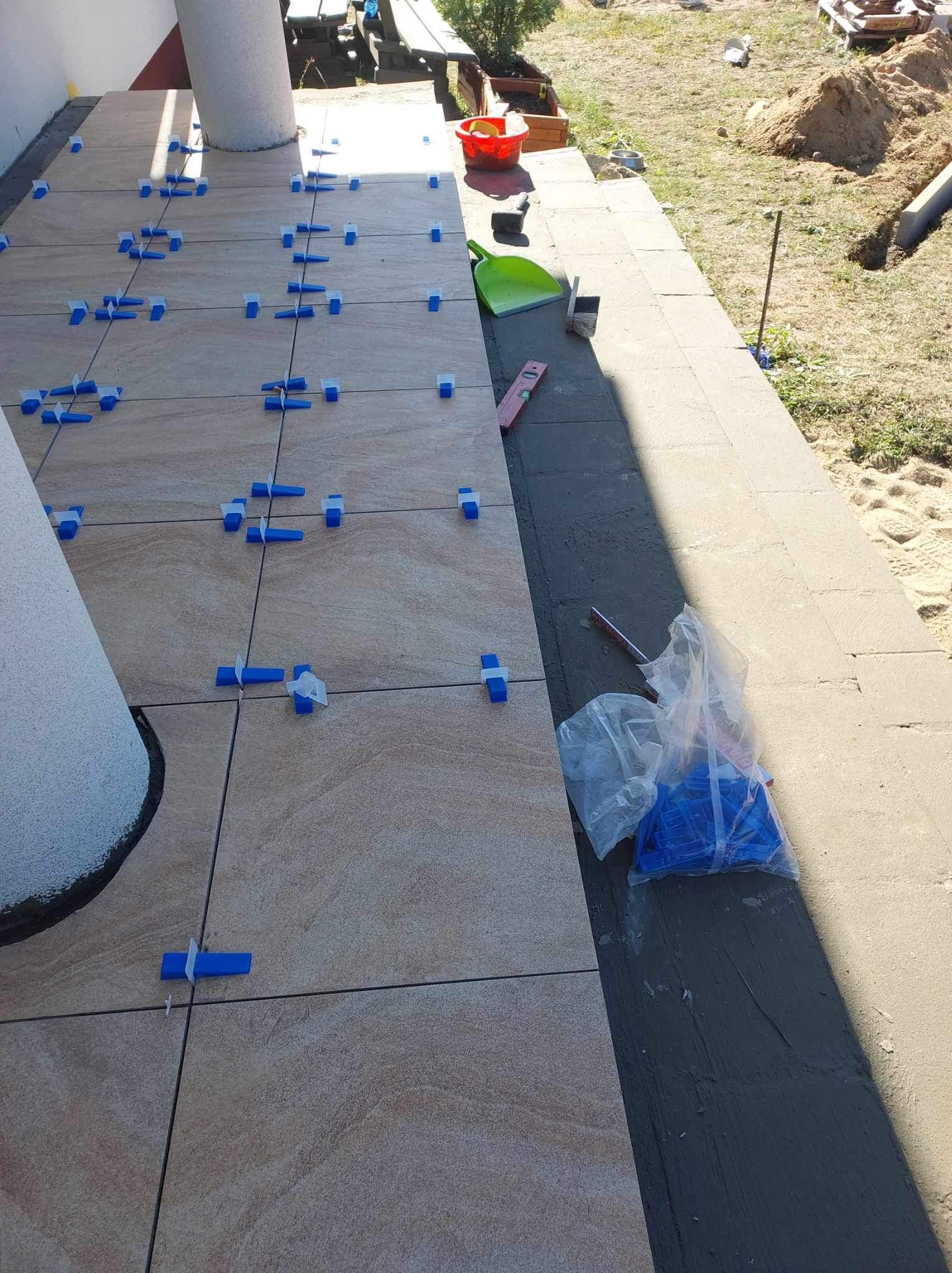 Układanie beżowych płytek na tarasie, widoczne niebieskie klipsy poziomujące, obok narzędzia i worki z klipsami, w tle kolumna budynku i trawnik.