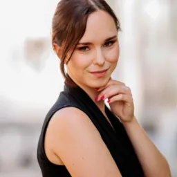 Portret uśmiechniętej kobiety w czarnej sukience, opierającej podbródek na dłoni z jaskraworóżowym manicure