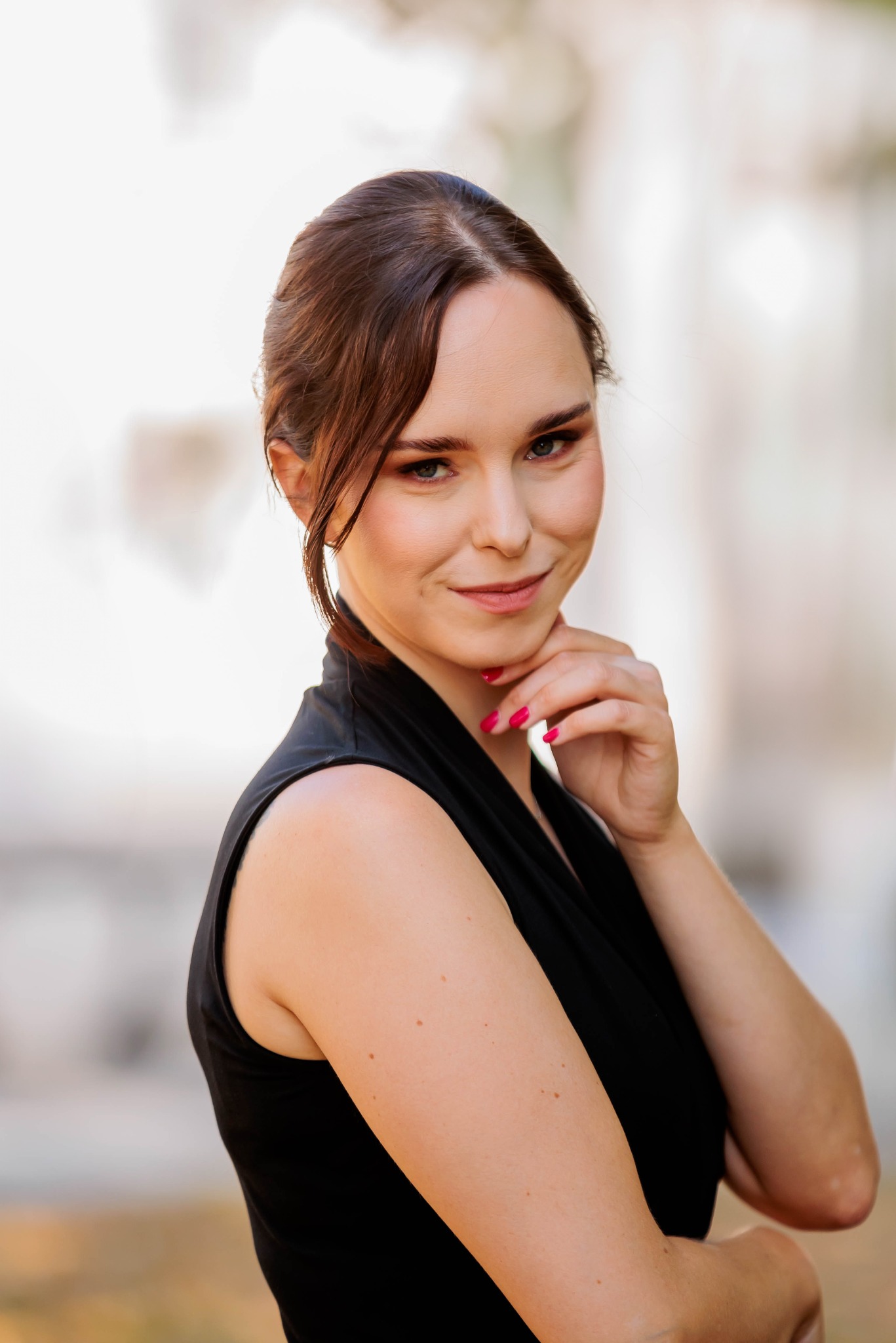 Portret uśmiechniętej kobiety w czarnej sukience, opierającej podbródek na dłoni z jaskraworóżowym manicure