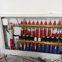 MAG-EKO - Rozdzielacz ogrzewania podłogowego w szafce, widoczne rury w izolacji czerwonej i niebieskiej, zawory z rotametrami, oraz elementy instalacyjne na podłodze.