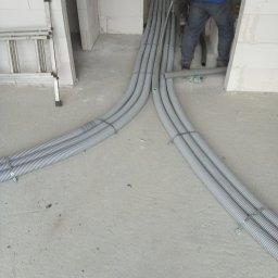 MAG-EKO - Szare, karbowane rury instalacyjne prowadzone po surowym betonowym podłożu w nowym budynku, rozdzielające się na dwie wiązki, widoczna drabina aluminiowa i fragment osoby w niebieskiej kurtce.