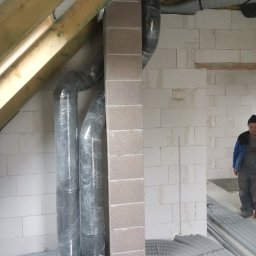 MAG-EKO - Wnętrze w trakcie montażu systemu wentylacji z widocznymi rurami wentylacyjnymi owiniętymi folią ochronną, ułożonymi na podłodze rurami oraz fragmentem ściany z betonu komórkowego i pracownikiem...