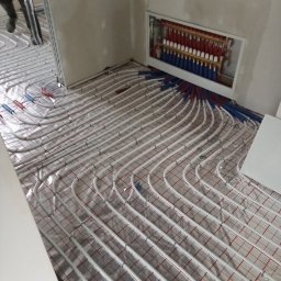 MAG-EKO - Rozłożone białe rury systemu ogrzewania podłogowego na folii z czerwoną siatką, widoczny rozdzielacz w ścianie i fragment osoby instalującej.