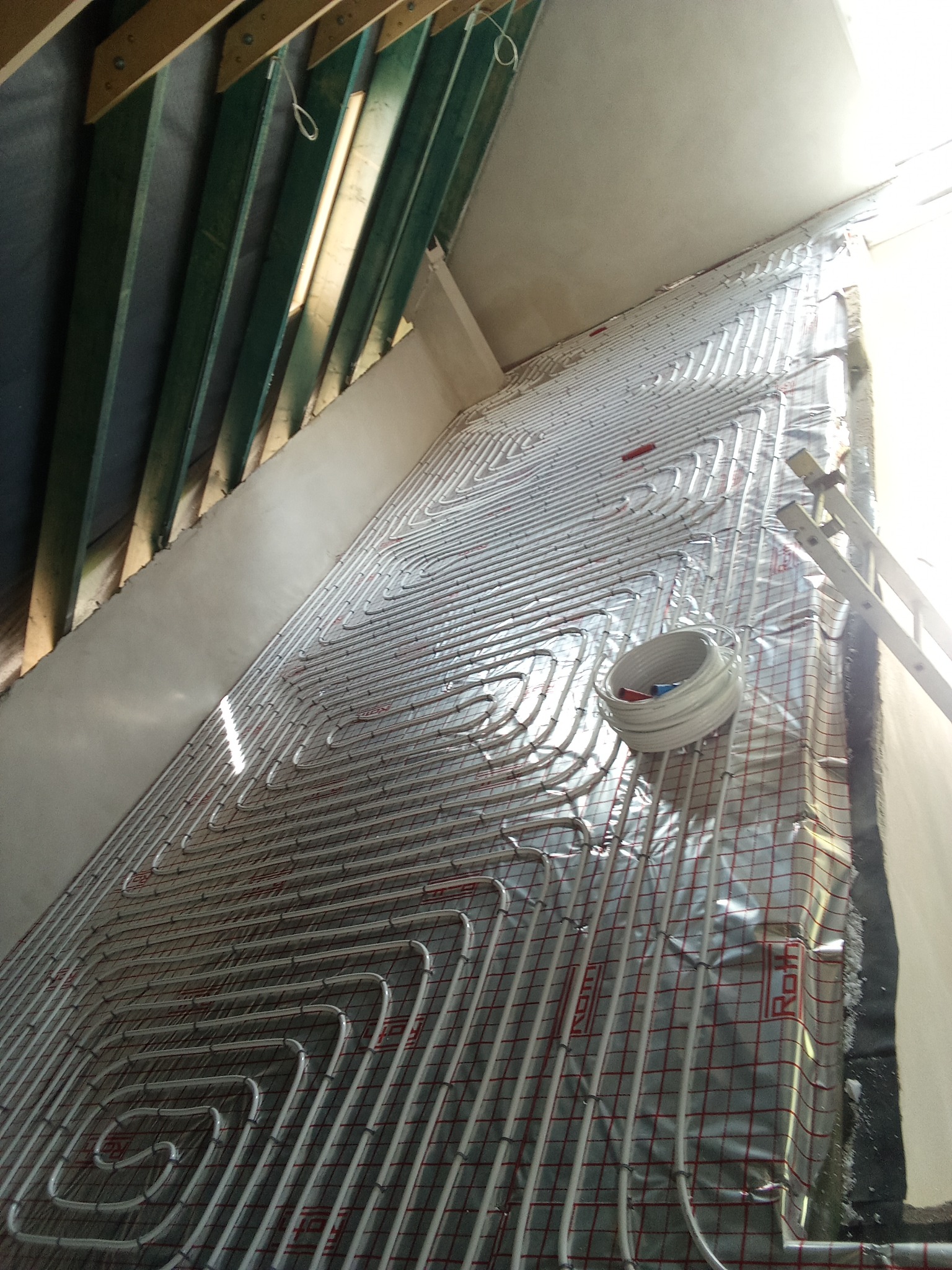 Białe rury układane spiralnie na folii z czerwoną siatką, prawdopodobnie system ogrzewania podłogowego, widoczne drewniane elementy konstrukcyjne dachu i drabina oparta o ścianę.