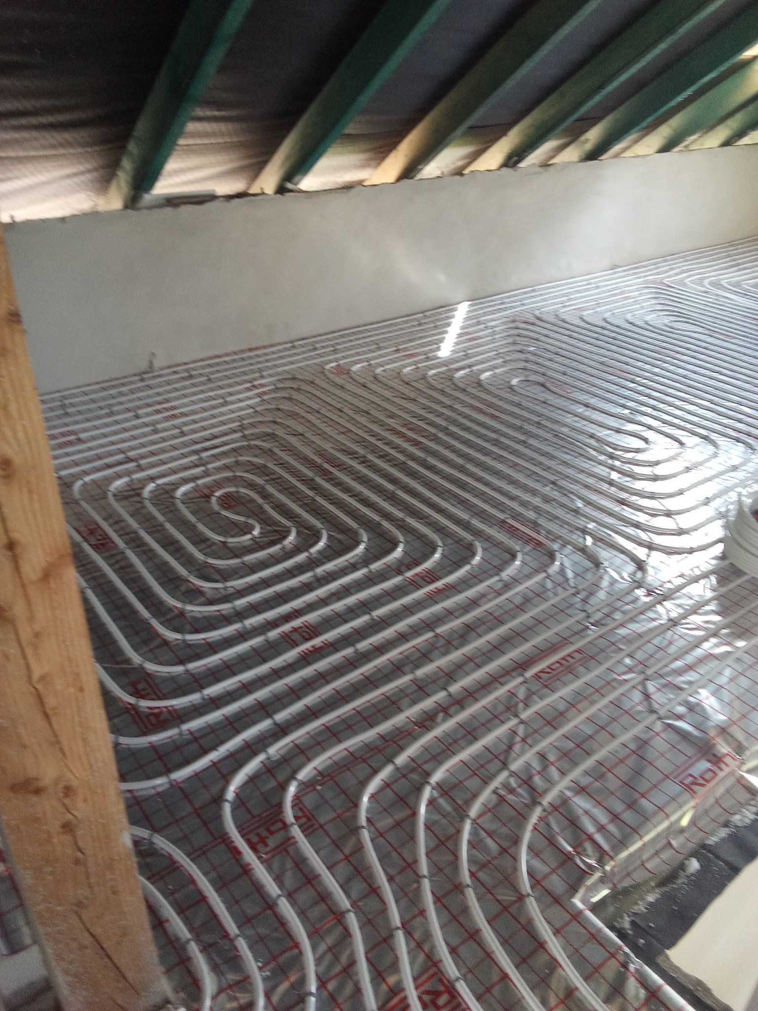 Białe rury systemu grzewczego podłogowego ułożone na folii z czerwoną siatką w surowym wnętrzu z drewnianą belką i widoczną izolacją poddasza.