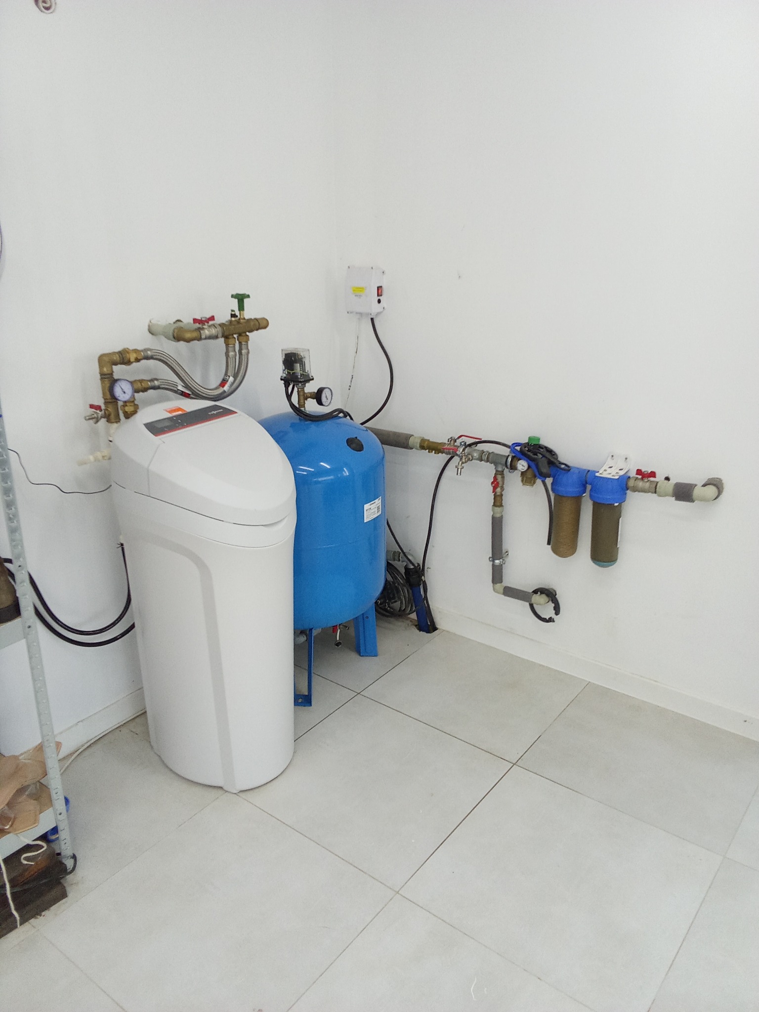 Zestaw filtrów do wody, w tym duży biały zmiękczacz wody i niebieski zbiornik ciśnieniowy, zamontowany w rogu pomieszczenia z białymi ścianami i jasną, kwadratową podłogą.