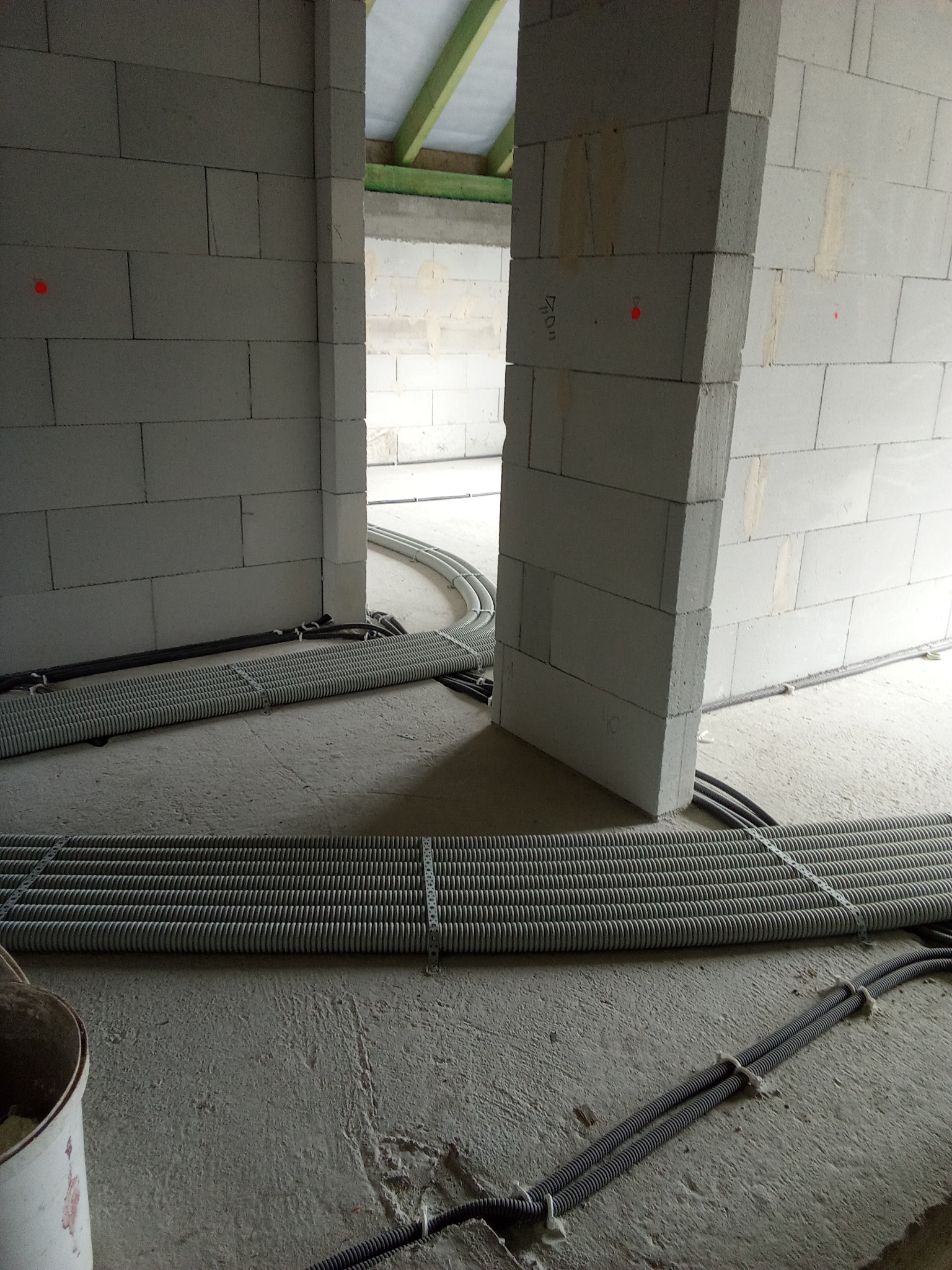 Wnętrze w trakcie budowy z widocznymi rurami wentylacyjnymi ułożonymi na podłodze w szarych peszlach, przymocowanych metalowymi obejmami, oraz fragmentem ściany z betonu komórkowego z zaznaczonymi...