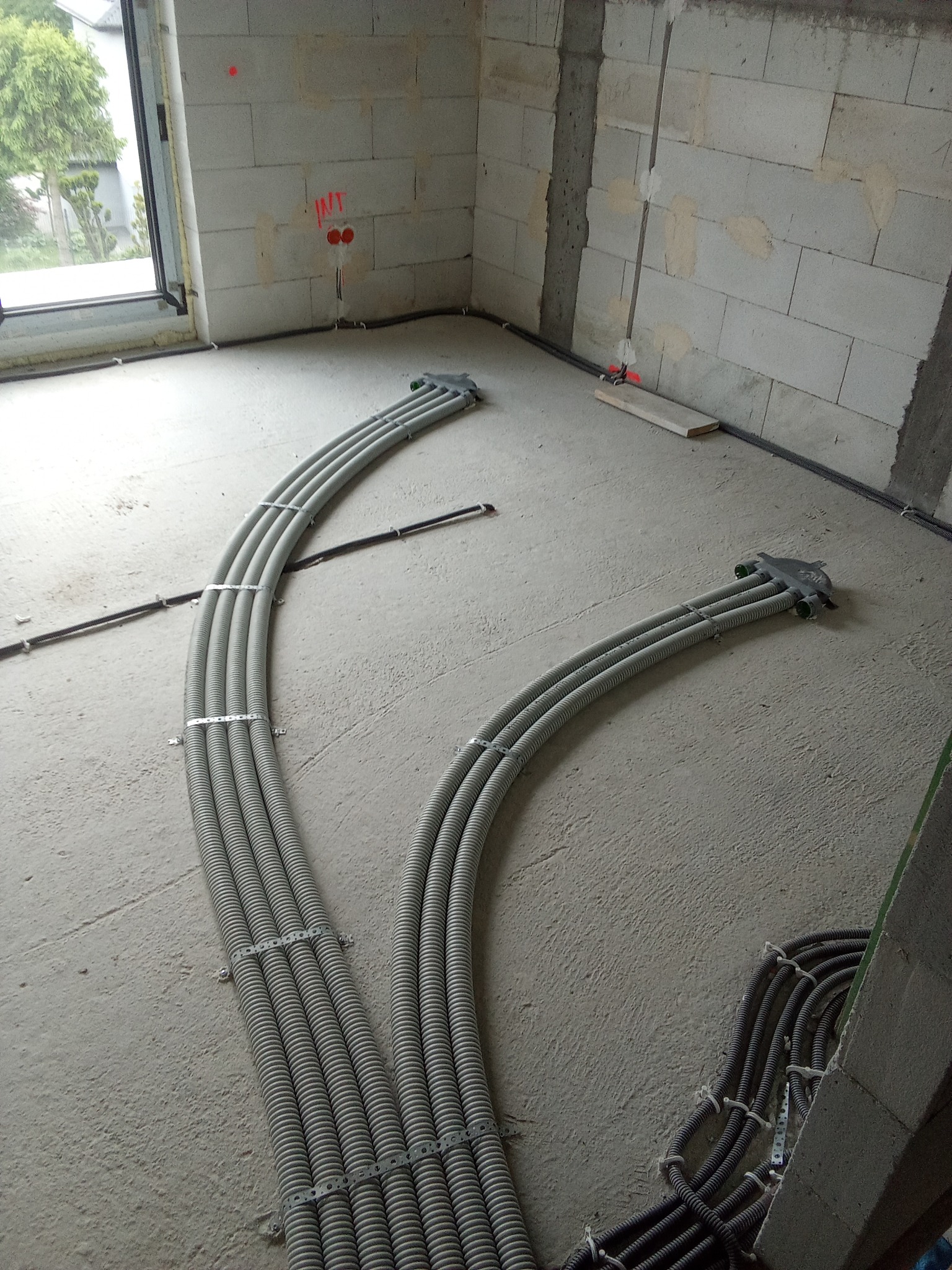 Szare, karbowane rury instalacyjne ułożone na surowym betonowym podłożu w nowym budynku, prowadzone do puszki rozdzielczej, z widocznymi mocowaniami i fragmentami czarnych kabli.