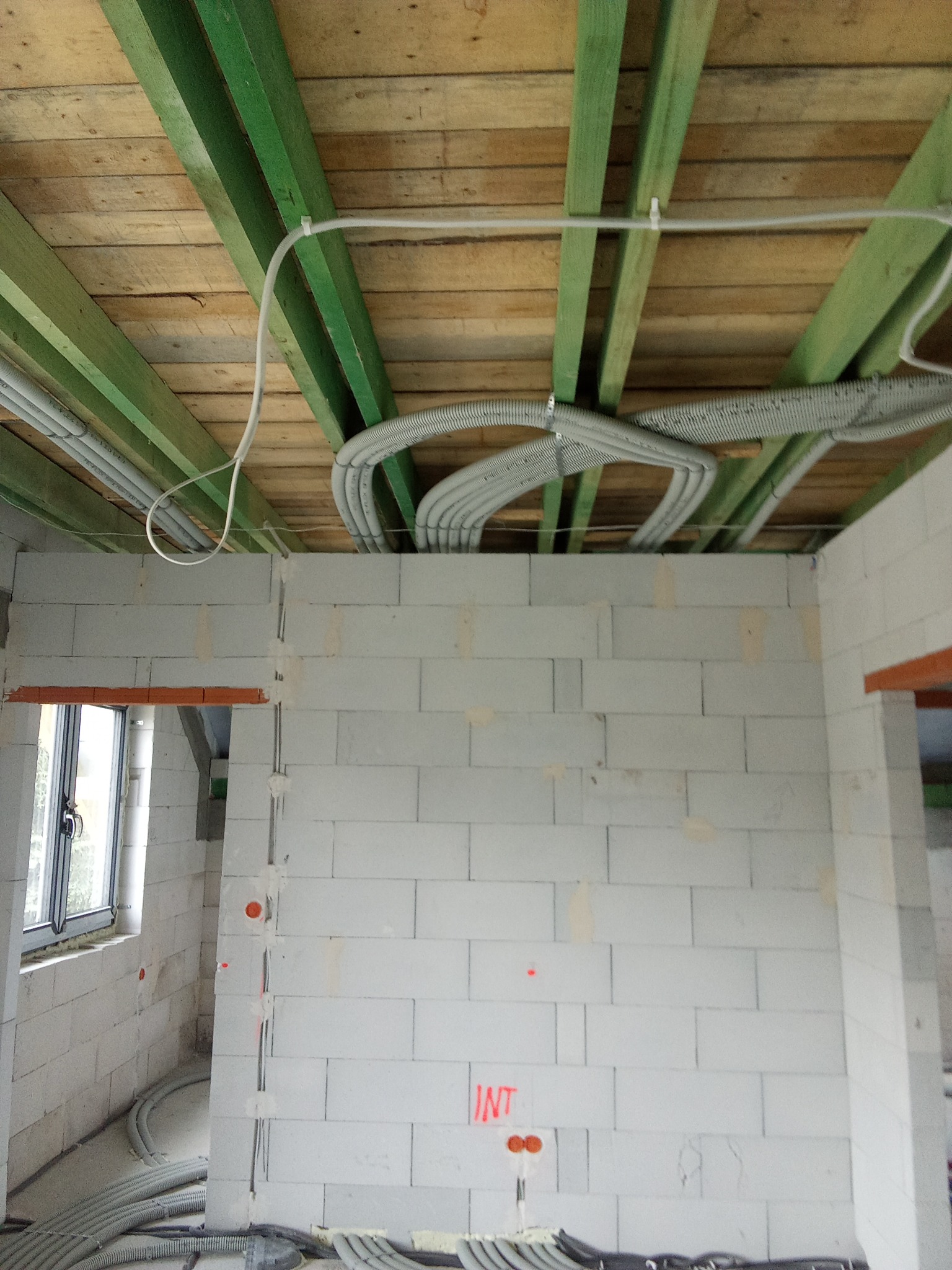 Surowe wnętrze w trakcie budowy z widocznymi przewodami wentylacyjnymi i elektrycznymi, poprowadzonymi pod drewnianym stropem i w ścianach z białych bloczków.