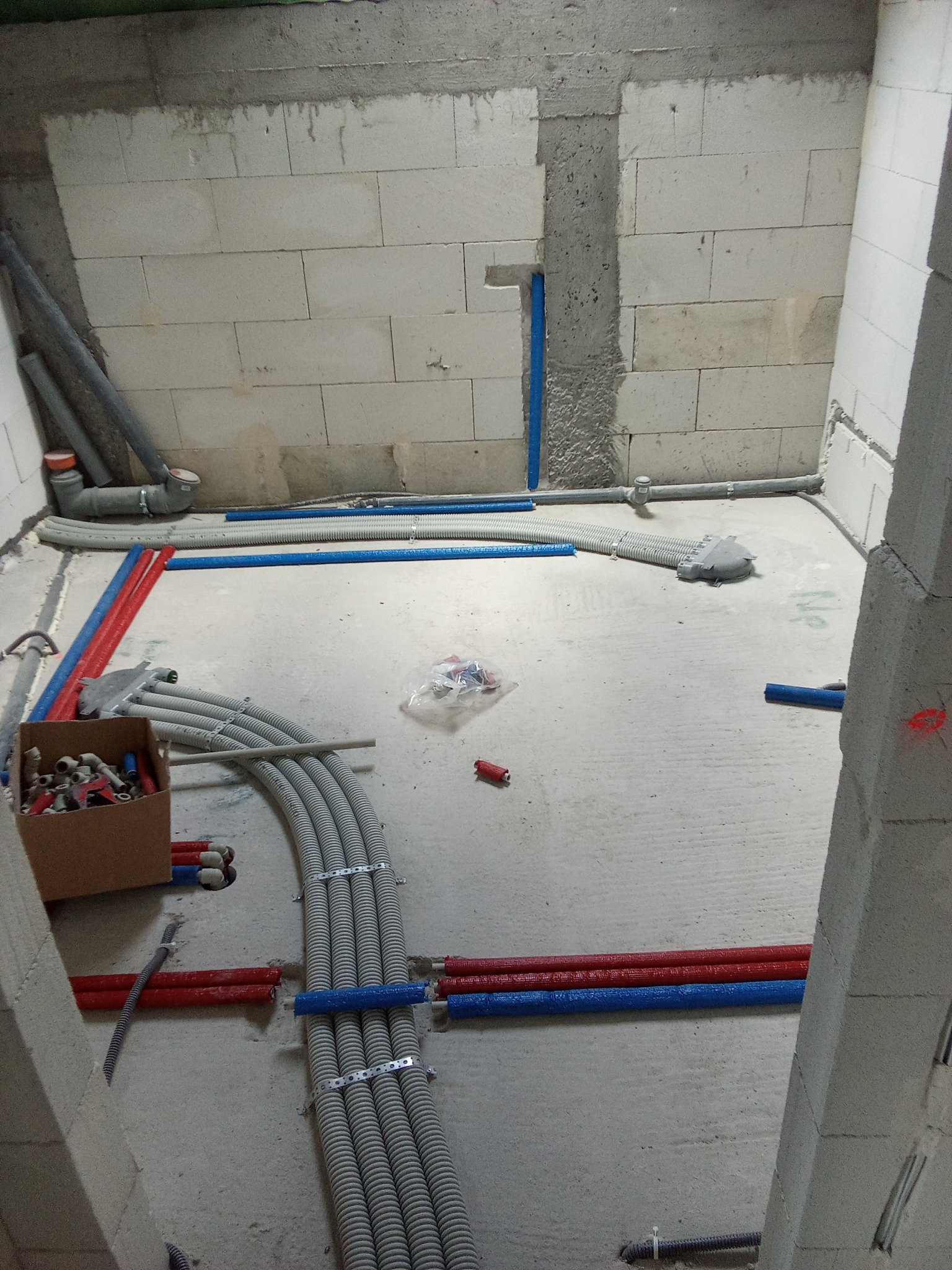Wnętrze w trakcie prac hydraulicznych: widoczne rury w kolorach czerwonym i niebieskim, szare rury karbowane mocowane do podłoża metalowymi klamrami, oraz karton złączek i kształtek w surowym...