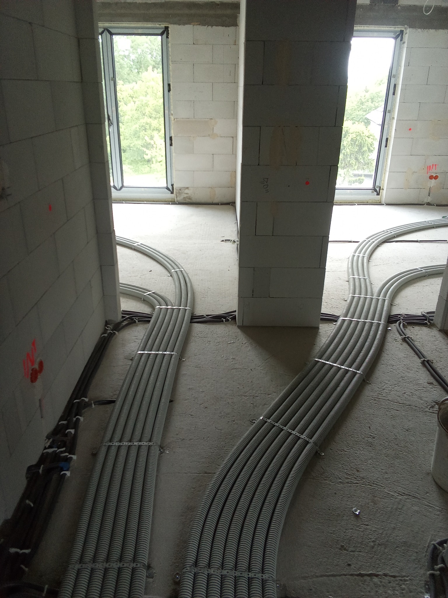 Szare rury instalacyjne ułożone wzdłuż ścian z betonu komórkowego w surowym stanie, prowadzone do otworów okiennych w nowym budynku.