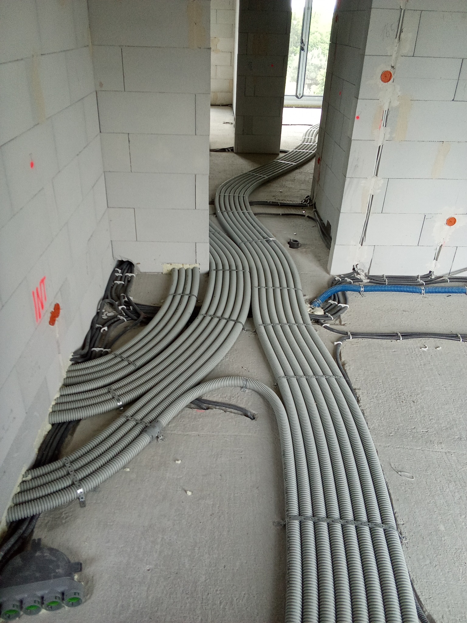 Szare rury karbowane prowadzone wzdłuż surowej podłogi w budynku w trakcie budowy, widoczne oznaczenia instalacyjne na ścianach z betonu komórkowego.