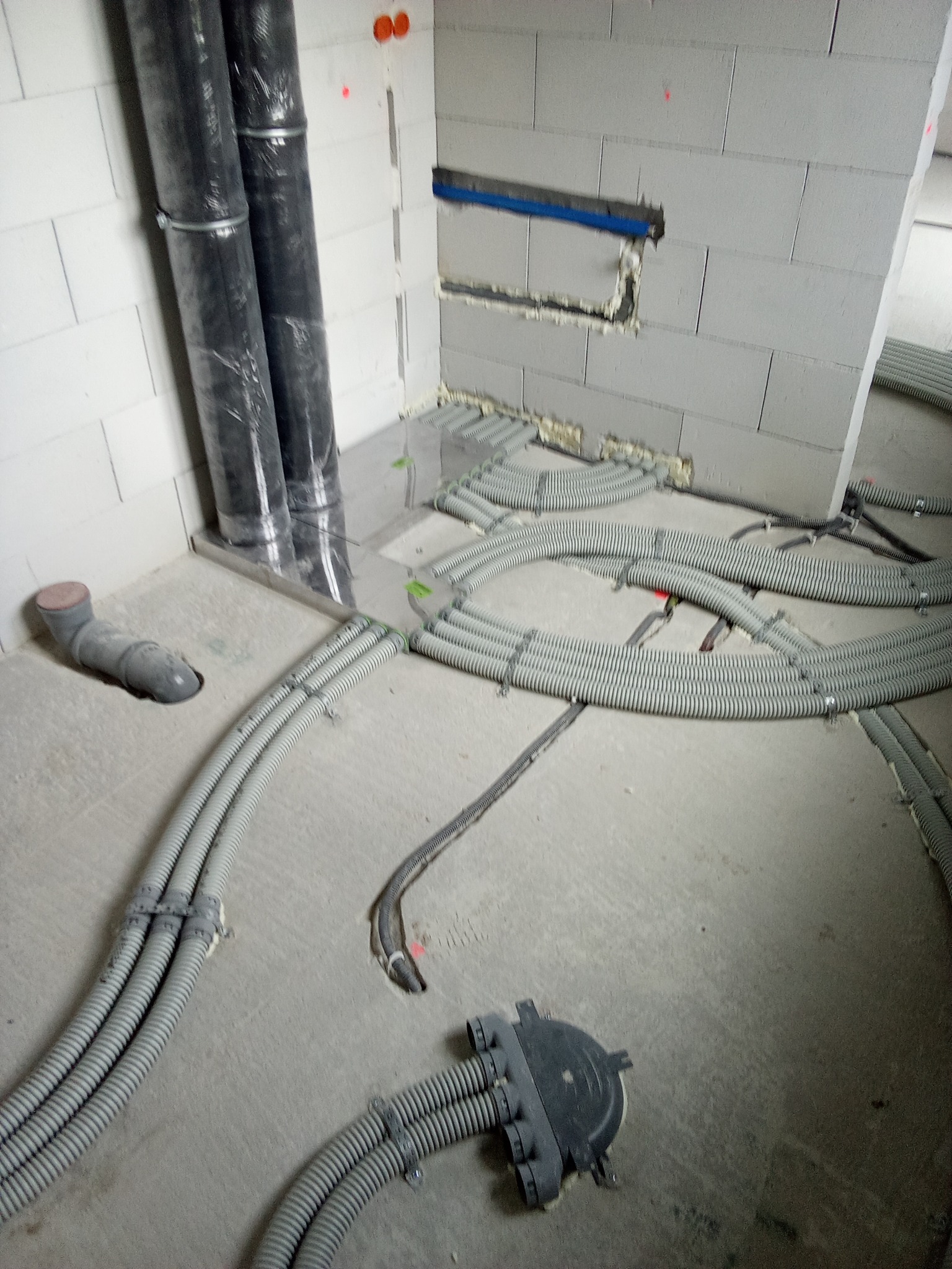 Szare, elastyczne rury wentylacyjne ułożone pętlami na surowej posadzce w trakcie instalacji, z widocznymi otworami w ścianie z betonu komórkowego i czarnymi rurami pionowymi w folii.