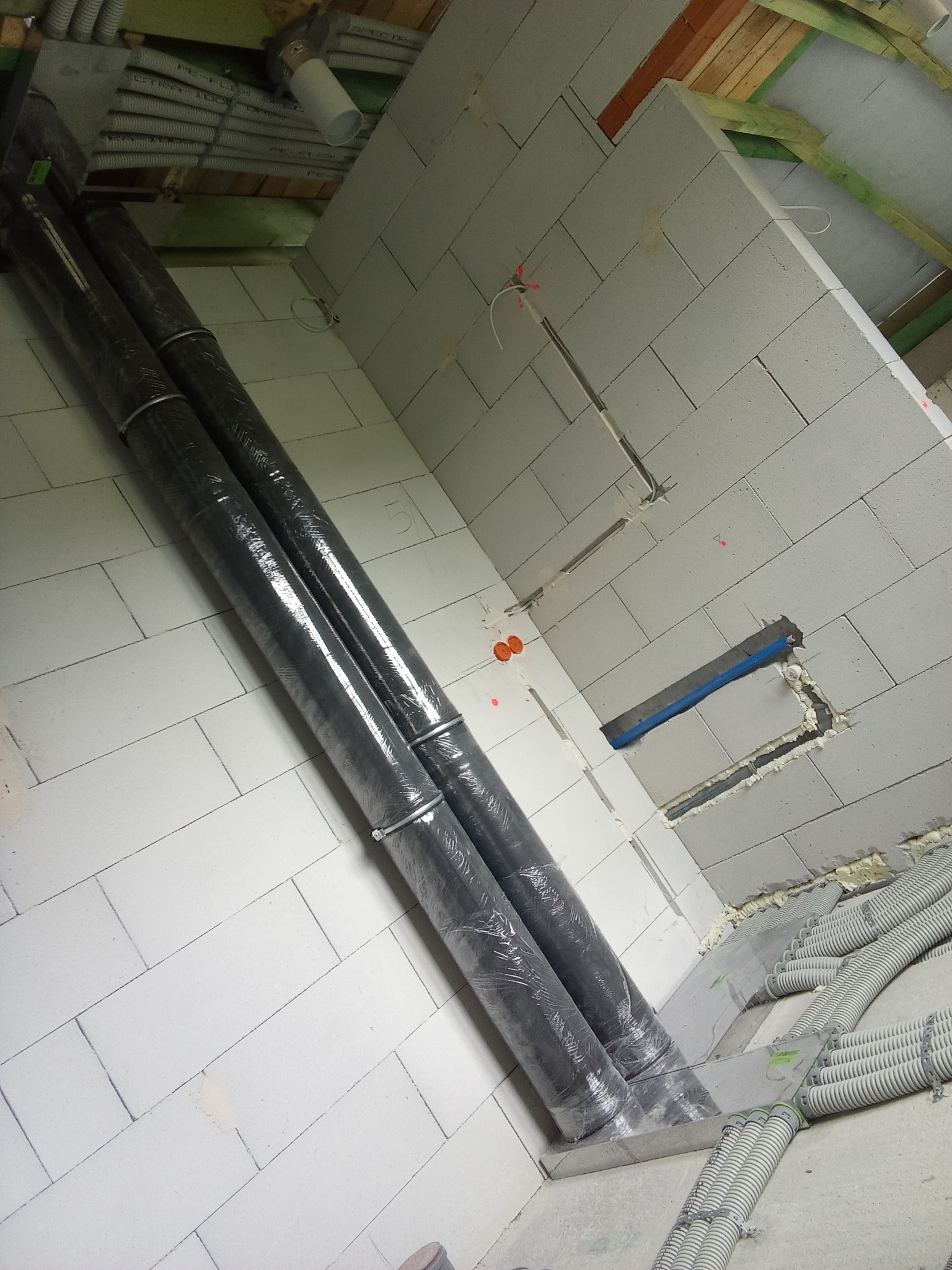Dwa rury wentylacyjne owinięte folią ochronną, zamocowane metalowymi obejmami, ułożone w surowym wnętrzu z białych bloczków, widoczne także peszle instalacyjne i fragmenty konstrukcji dachu.