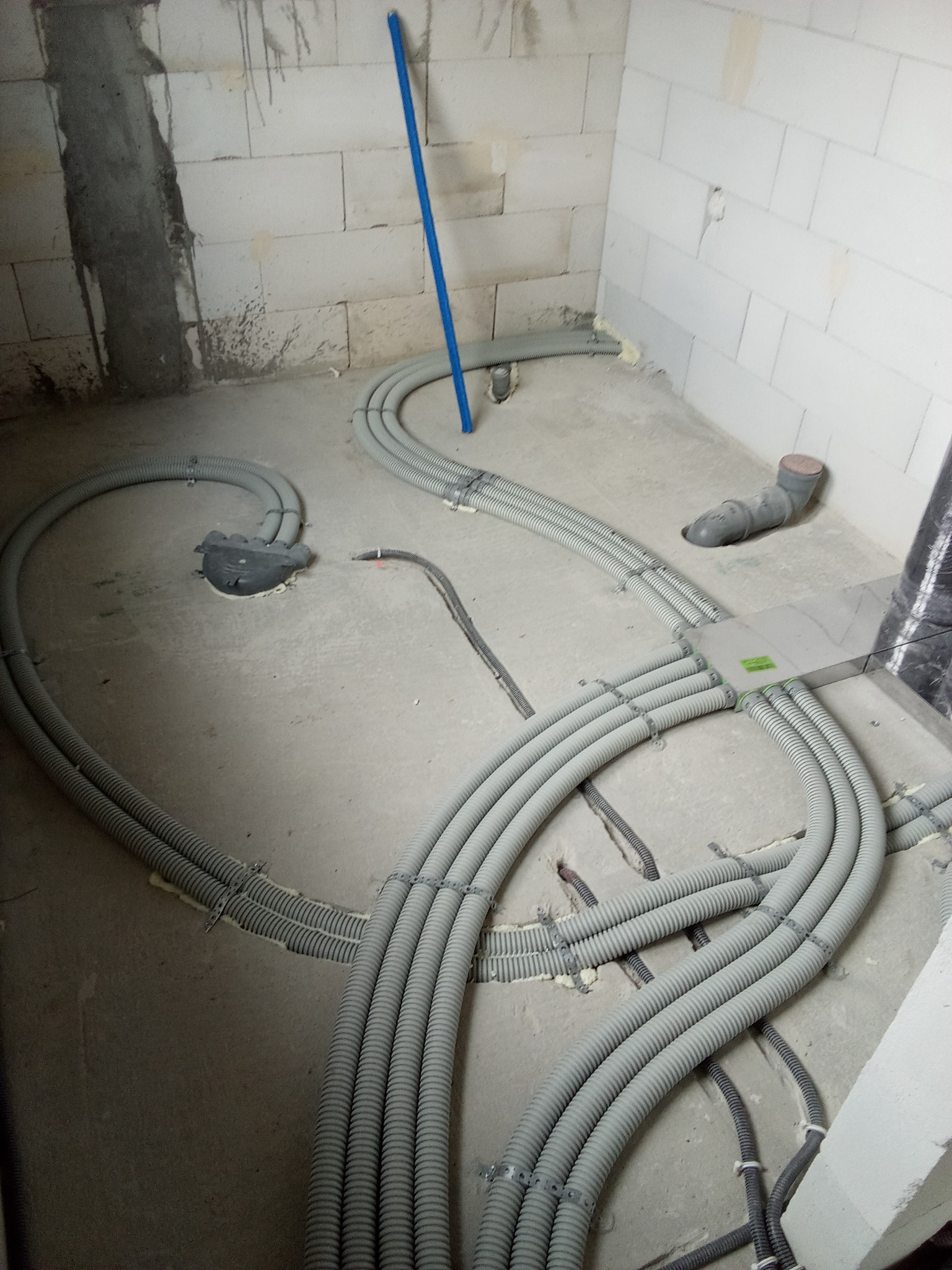 Szare, karbowane rury instalacyjne ułożone na surowym betonowym podłożu w trakcie montażu, prowadzące do rozdzielacza z zieloną naklejką, widoczne uchwyty mocujące rury do podłoża i fragmenty...