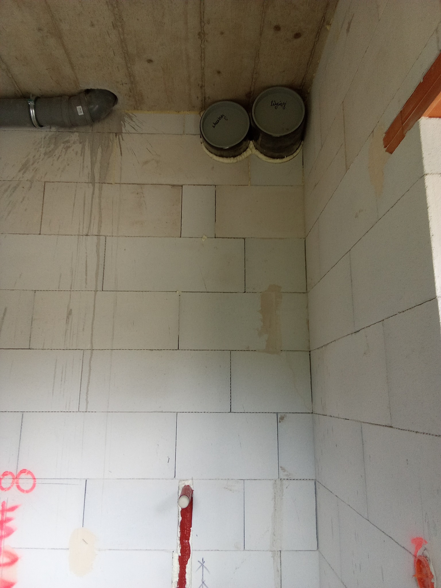 Fragment surowego wnętrza z widocznymi elementami instalacji: szary przewód wentylacyjny z metalową obejmą, dwa okrągłe otwory wentylacyjne w suficie zabezpieczone pianką montażową, oraz pionowy...