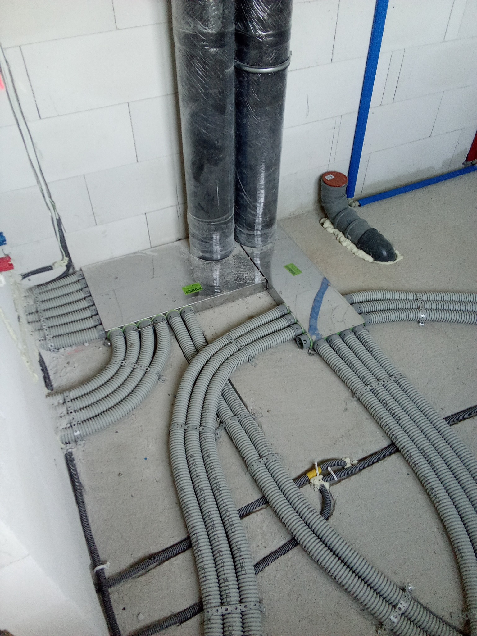 Szare rury wentylacyjne karbowane, częściowo schowane pod metalową płytą, prowadzone po surowym betonowym podłożu wzdłuż ściany z białych bloczków.