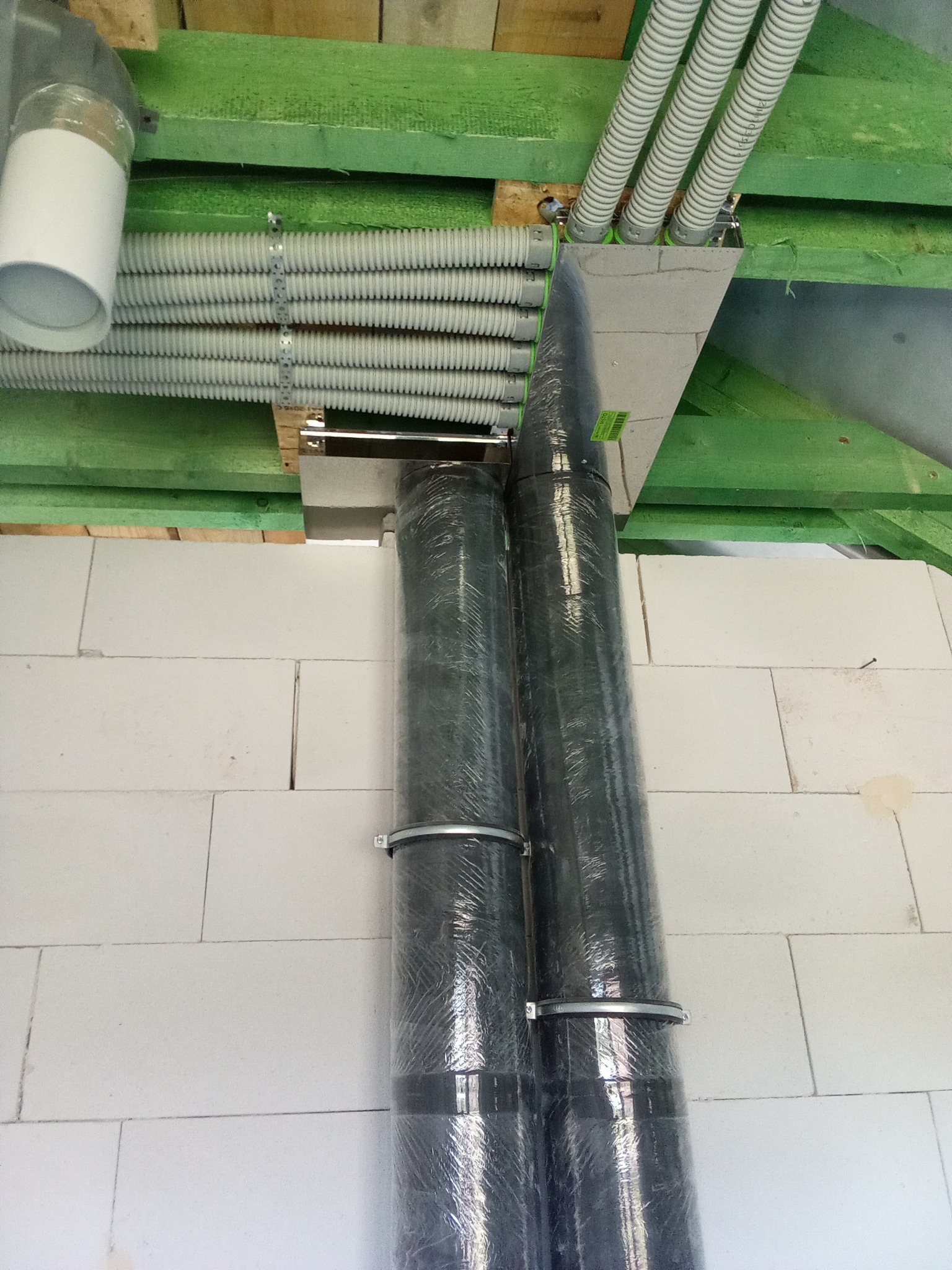 Dwa pionowe przewody w folii ochronnej, zamocowane do ściany z białych bloczków, z widocznymi poziomymi rurami wentylacyjnymi pod stropem z zielonych belek.