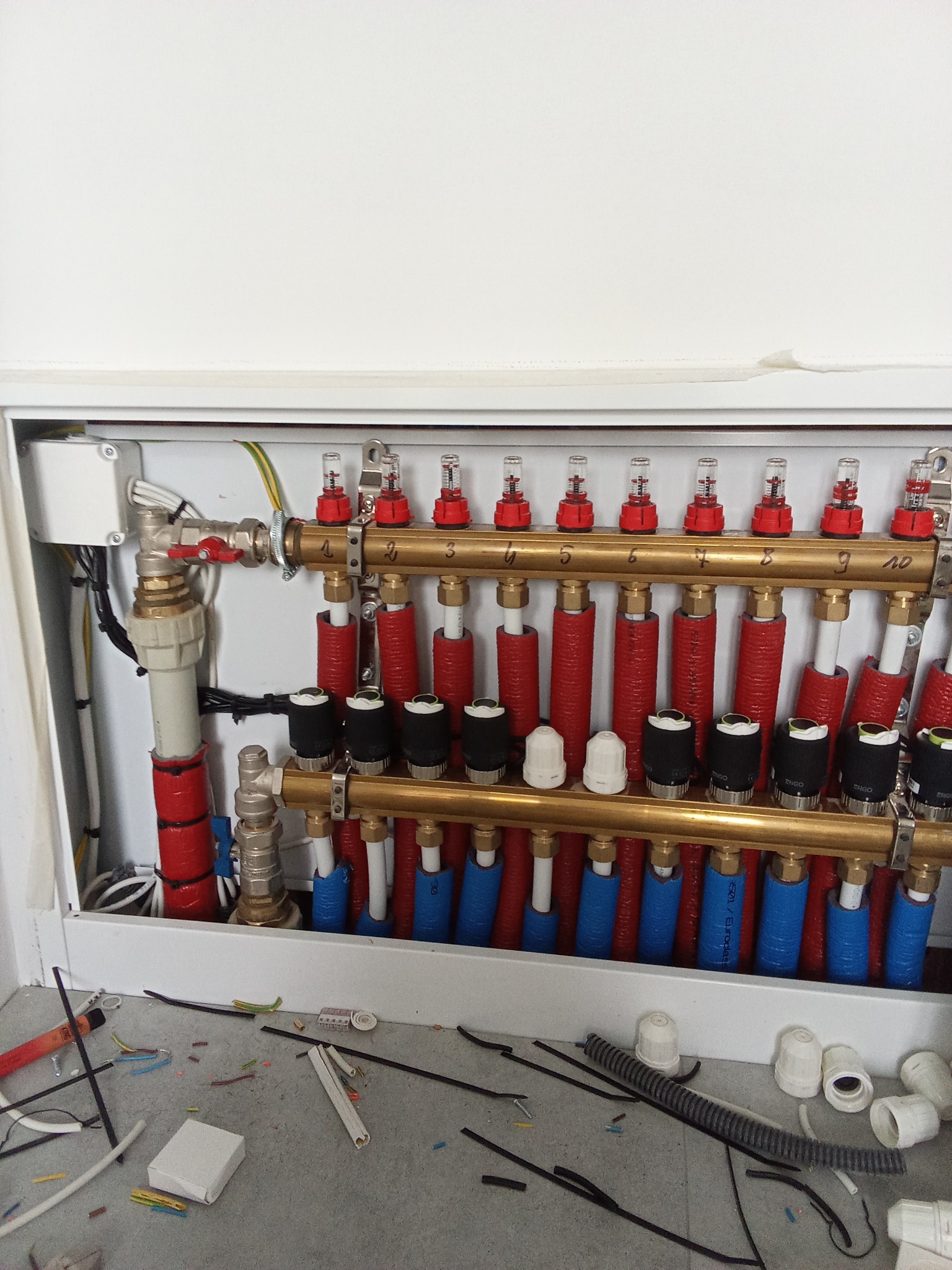 Rozdzielacz ogrzewania podłogowego w szafce, widoczne rury w izolacji czerwonej i niebieskiej, zawory z rotametrami, oraz elementy instalacyjne na podłodze.