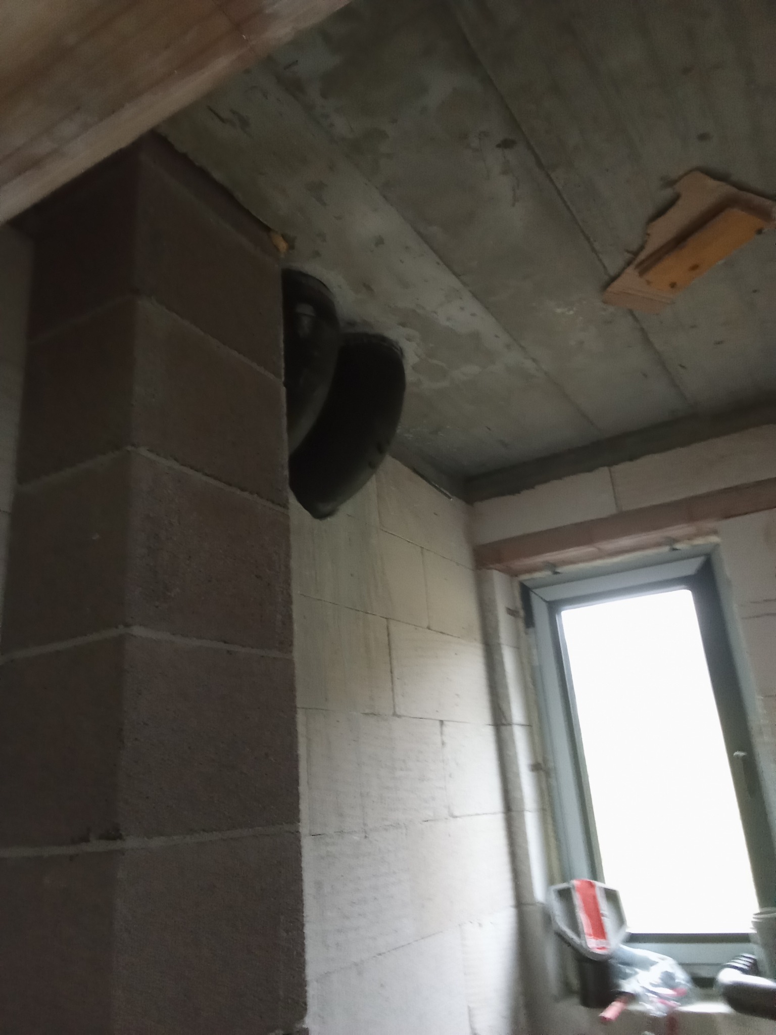 Fragment instalacji wentylacyjnej z dwoma czarnymi kanałami wentylacyjnymi, prowadzonej w rogu surowego pomieszczenia z oknem, widoczne bloczki betonowe i surowy betonowy strop.