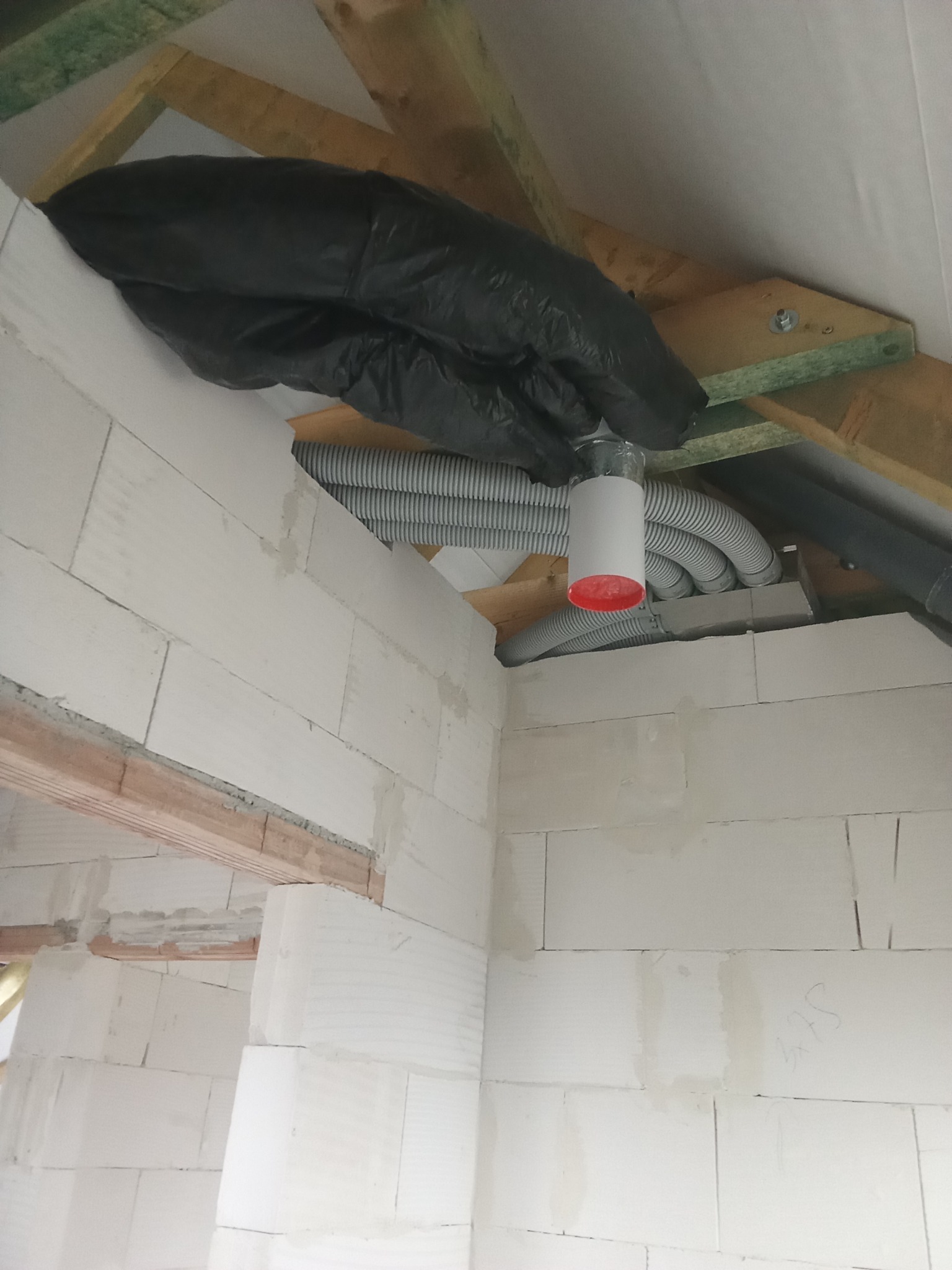 Szare, elastyczne rury wentylacyjne pod stropem, częściowo osłonięte czarną folią, widoczne elementy konstrukcji drewnianej i ściany z betonu komórkowego w surowym stanie.