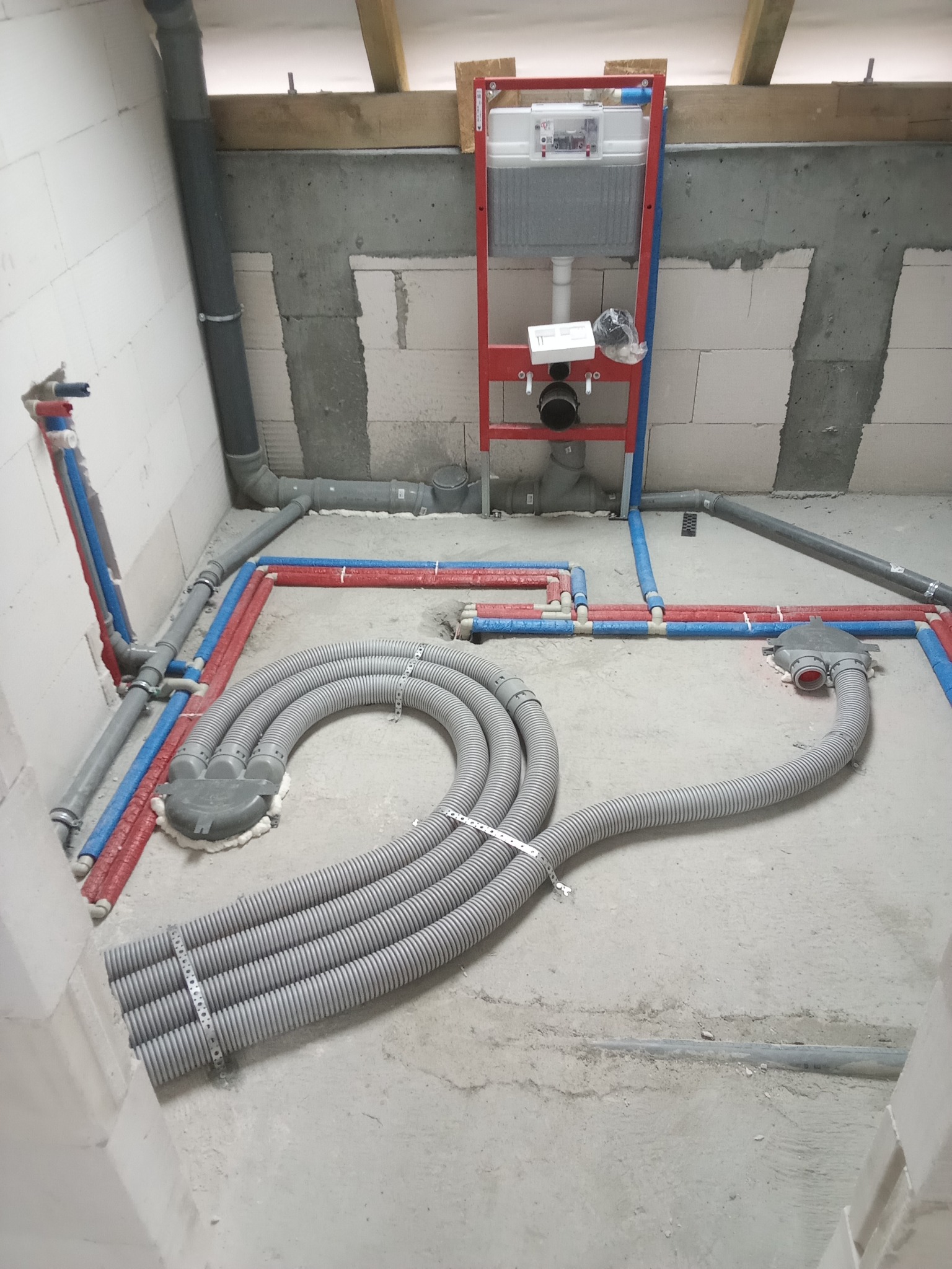 Surowy stan łazienki w trakcie prac hydraulicznych z widocznym stelażem podtynkowym WC, rurami doprowadzającymi wodę, szarymi rurami kanalizacyjnymi i czterema szarymi elastycznymi przewodami...