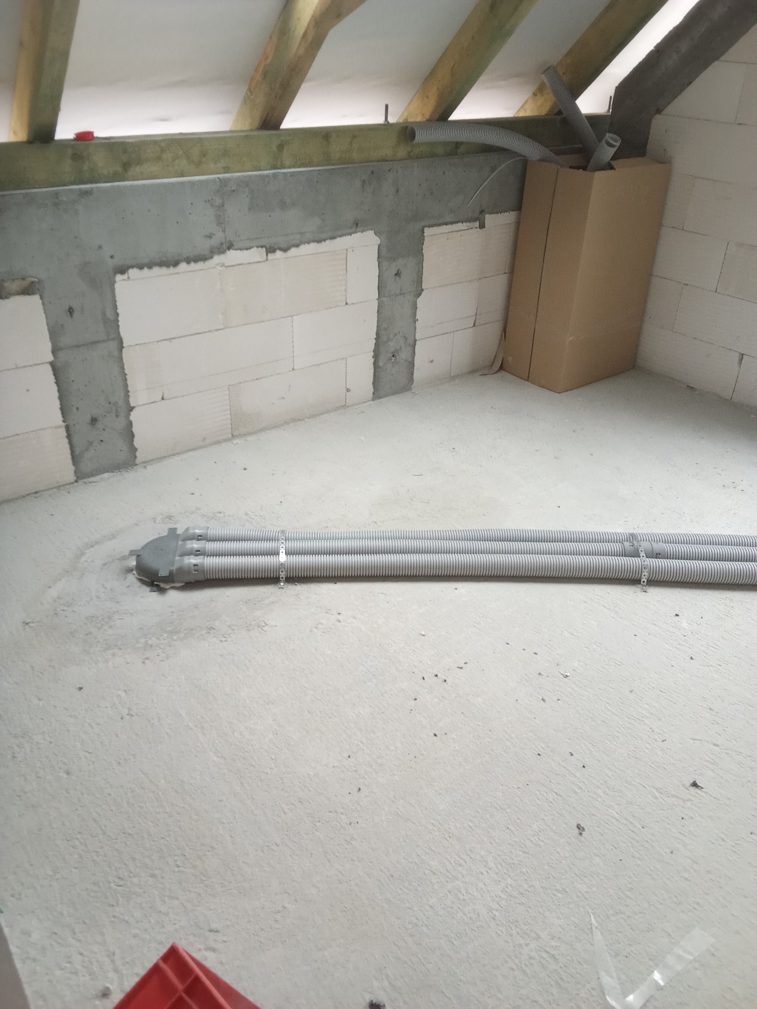 Trzy elastyczne rury wentylacyjne w szarej osłonie wychodzące z podłoża w surowym stanie, widoczne fragmenty ścian z betonu komórkowego i drewniana konstrukcja dachu na poddaszu.