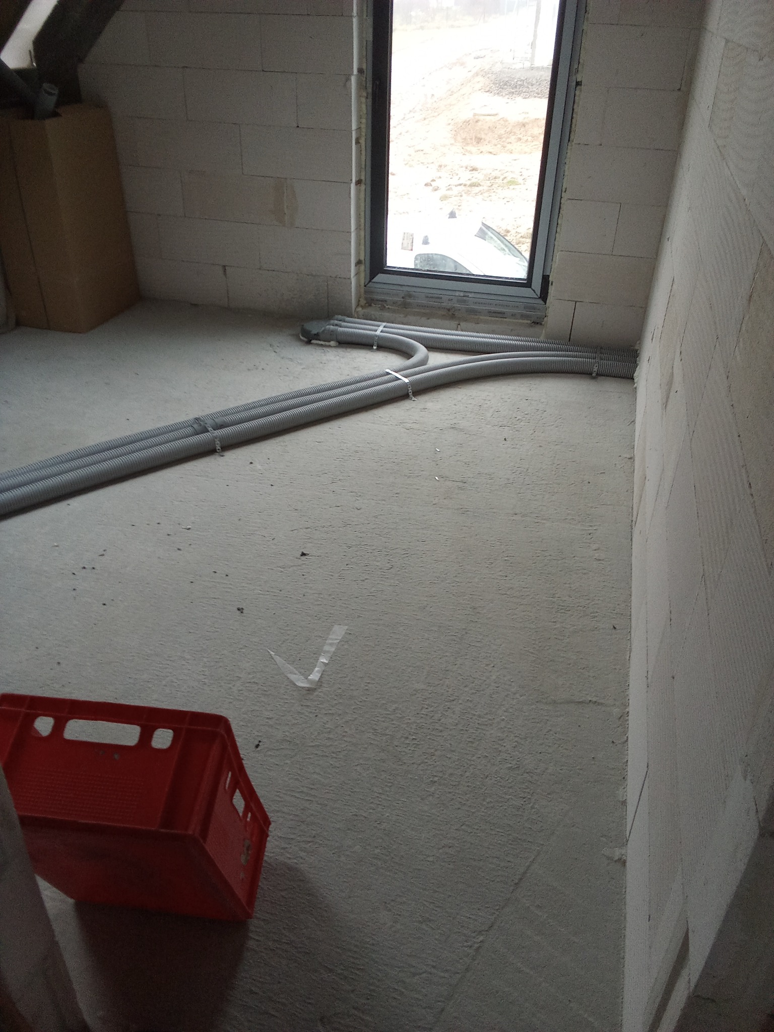 Wstępny etap instalacji wentylacji w Wiśniowej: widok na trzy elastyczne rury wentylacyjne prowadzące do okna w surowym pomieszczeniu z czerwoną skrzynką w rogu.