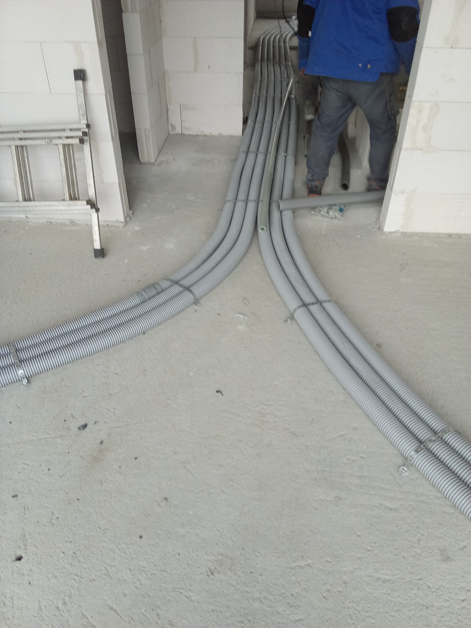 Szare, karbowane rury instalacyjne prowadzone po surowym betonowym podłożu w nowym budynku, rozdzielające się na dwie wiązki, widoczna drabina aluminiowa i fragment osoby w niebieskiej kurtce.