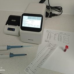 Gabinet Weterynaryjny Centaur - Analizator biochemiczny Mncchip z ekranem, drukarka etykiet, dwie pipety, wydruk z wynikami badań i probówka z krwią na blacie.