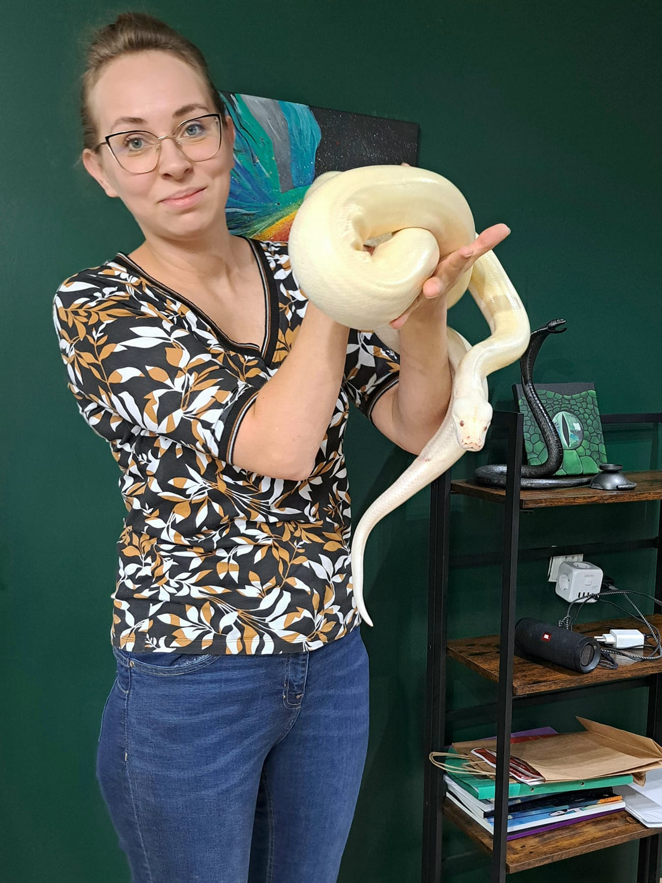 Kobieta w okularach, trzymająca w dłoniach dużego, białego węża albinosa, z półką w tle.