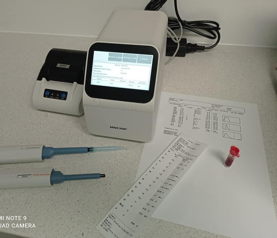 Analizator biochemiczny Mncchip z ekranem, drukarka etykiet, dwie pipety, wydruk z wynikami badań i probówka z krwią na blacie.