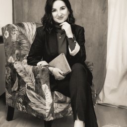 Sewastynowicz Czułe Wnętrze - Gabinet coachingowo - terapeutyczny - Portret kobiety w ciemnym garniturze siedzącej w fotelu z motywem roślinnym, trzymającej w ręku książkę i uśmiechającej się, w tle zasłona.