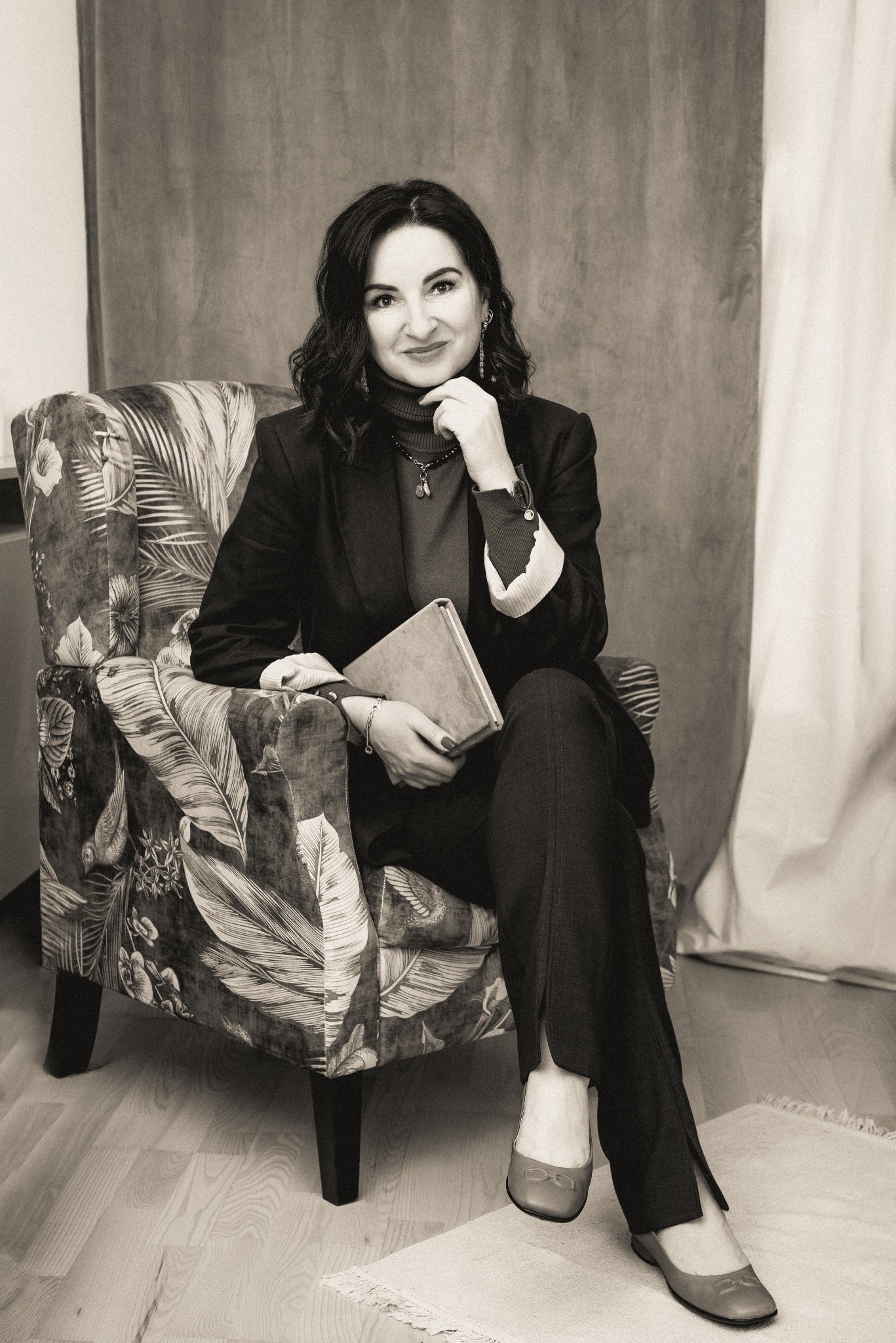 Portret kobiety w ciemnym garniturze siedzącej w fotelu z motywem roślinnym, trzymającej w ręku książkę i uśmiechającej się, w tle zasłona.