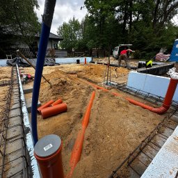 Ultra Service Oleksandr Harhalyk - Instalacja rur kanalizacyjnych w wykopie fundamentowym, widoczne zbrojenie, izolacja termiczna i pracownik wyrównujący teren.