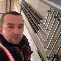 Ultra Service Oleksandr Harhalyk - Instalator pozuje na tle pionowo ułożonych rur grzewczych, część z nich zaizolowana czarną pianką, mocowanych do ściany za pomocą metalowych uchwytów.