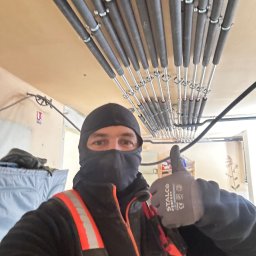 Ultra Service Oleksandr Harhalyk - Instalator w kominiarce i odblaskowej kamizelce, z kciukiem w górę, prezentuje rury grzewcze zamontowane pod sufitem w Warszawie. Rury są zabezpieczone obejmami.