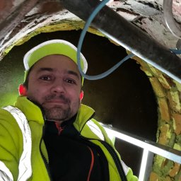Ultra Service Oleksandr Harhalyk - Instalator w odblaskowej kurtce i czapce, robiący selfie wewnątrz ceglanego kanału z rurami i oświetleniem, widok z dołu.