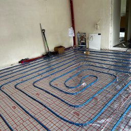 Ultra Service Oleksandr Harhalyk - Niebieskie rury ogrzewania podłogowego rozłożone na folii z czerwoną siatką, widoczna rozdzielnia i narzędzia w surowym wnętrzu w trakcie prac instalacyjnych.