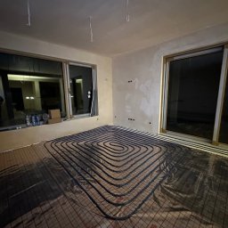 Ultra Service Oleksandr Harhalyk - Wnętrze w trakcie prac: rozłożone rury ogrzewania podłogowego na folii z siatką, widoczne okna przesuwne i przygotowane ściany do wykończenia.