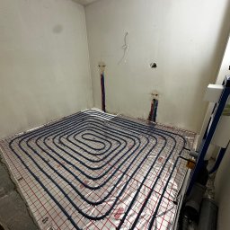 Ultra Service Oleksandr Harhalyk - Niedokończona instalacja ogrzewania podłogowego w surowym pomieszczeniu, widoczne rury układane na folii z siatką, wystające rury instalacyjne w ścianie, brak wykończenia ścian.