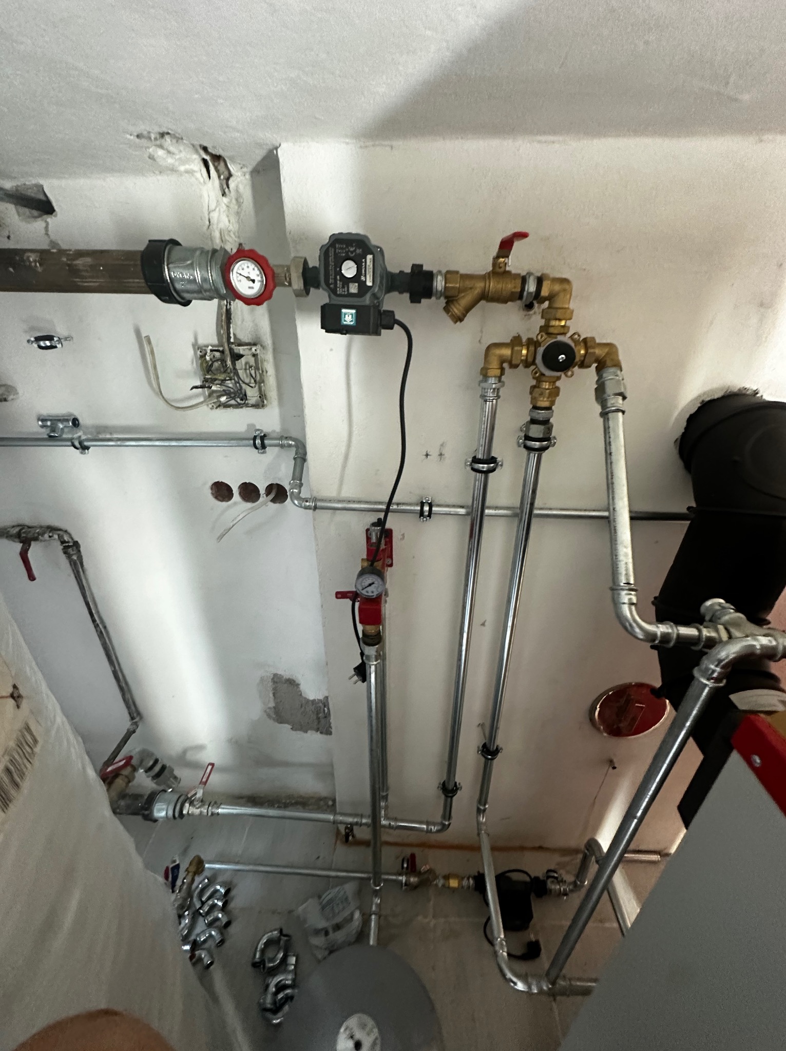Widok instalacji hydraulicznej i grzewczej pod sufitem w trakcie montażu, widoczne rury stalowe, mosiężne zawory, manometr, pompa obiegowa,  otwory w ścianie i przewody elektryczne bez puszek...