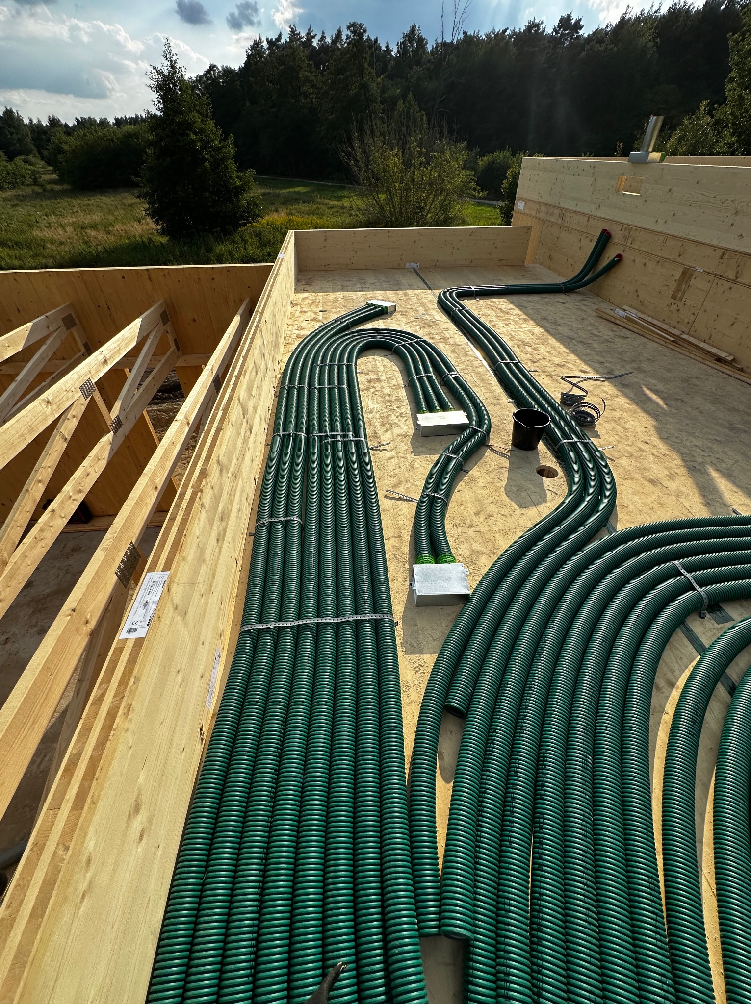 Zielone rury wentylacyjne ułożone na drewnianej konstrukcji dachu w trakcie budowy, widoczne elementy mocujące i skrzynki przyłączeniowe, w tle krajobraz z drzewami.