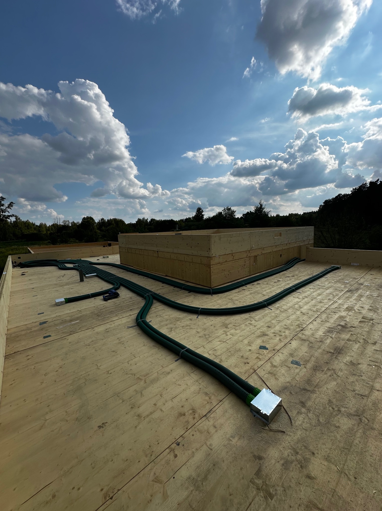 Zielone rury wentylacyjne rozłożone na drewnianej konstrukcji stropu z widocznym niebem i drzewami w tle.