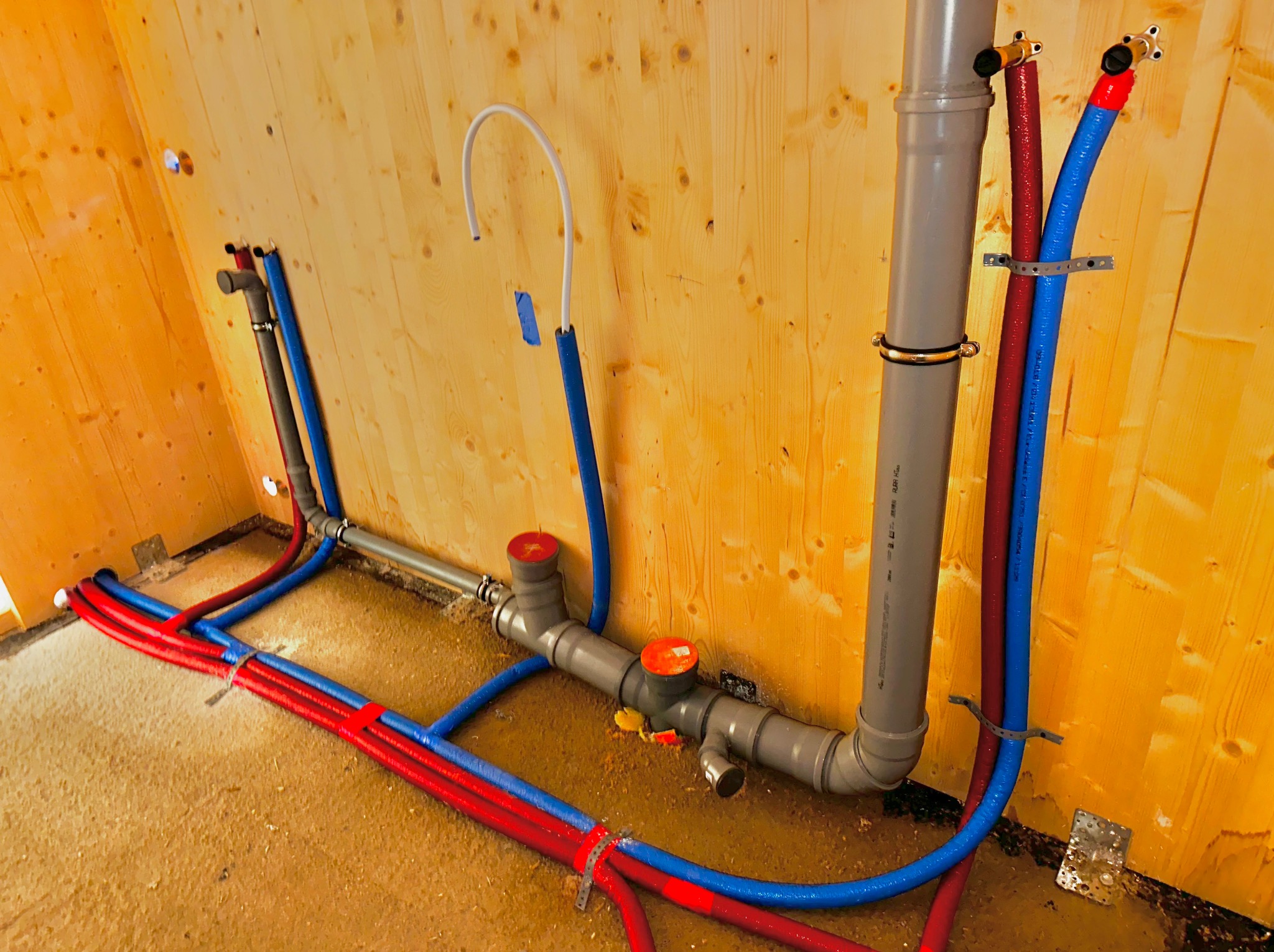Wewnętrzne instalacje wodne i kanalizacyjne w trakcie montażu na drewnianej ścianie, widoczne rury PEX w kolorach czerwonym i niebieskim, szare rury kanalizacyjne z pomarańczowymi zaślepkami,...