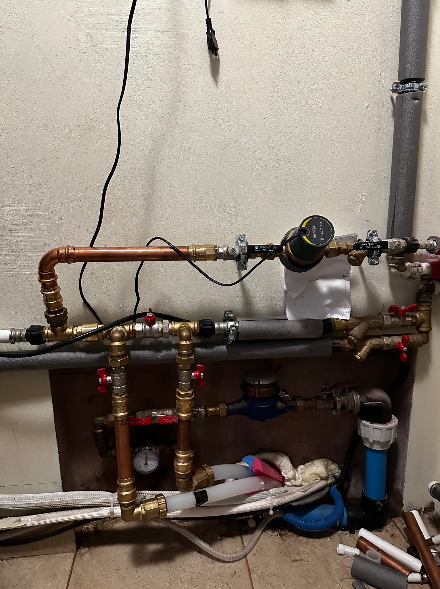 Skomplikowana instalacja hydrauliczna i grzewcza z miedzianymi rurami, zaworami, pompą obiegową i licznikiem, widoczna na tle ściany i podłogi w trakcie prac montażowych, z widocznymi elementami...