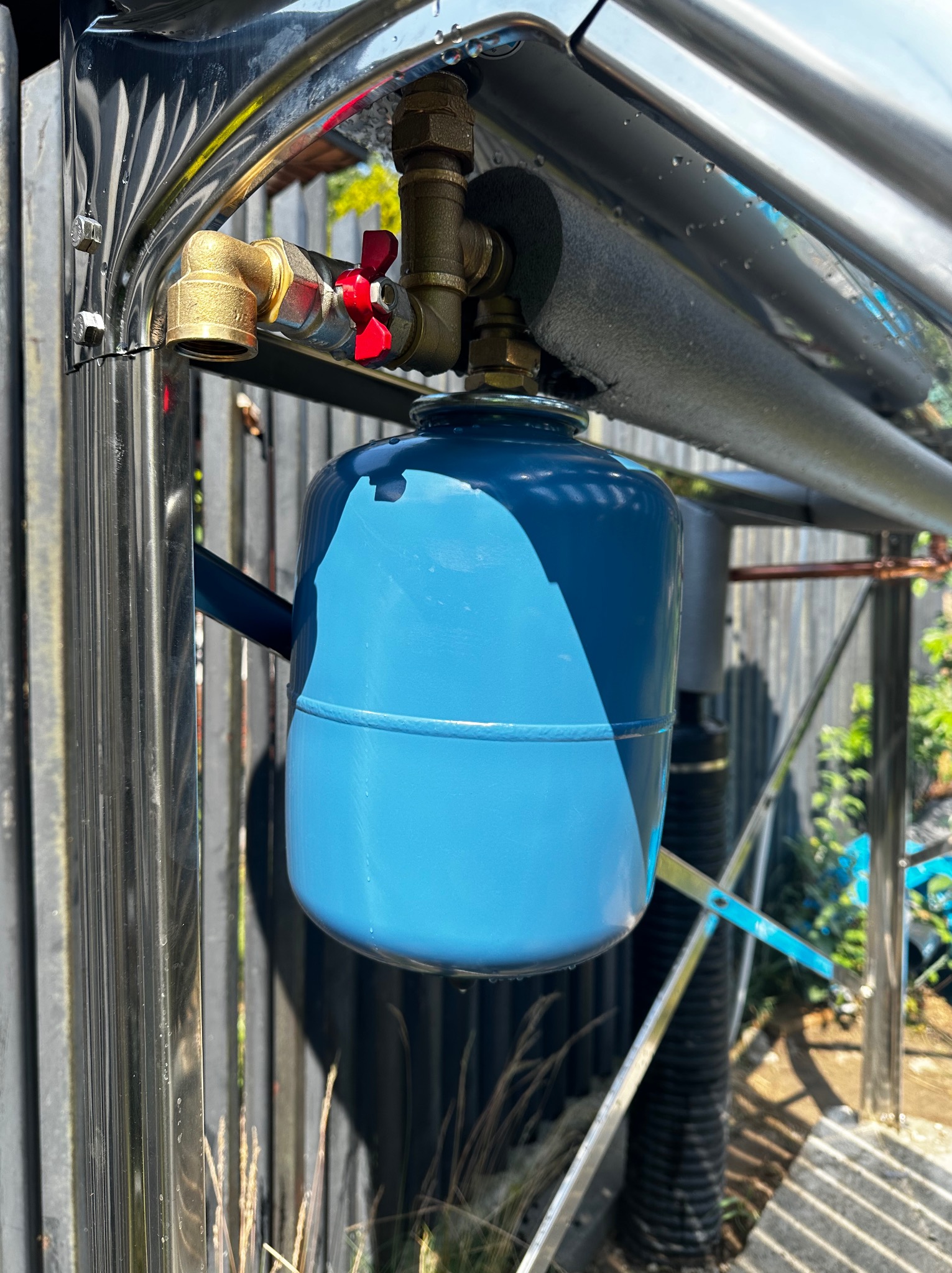 Niebieski zbiornik wyrównawczy w instalacji solarnej, widoczne rury chromowane, zawór z czerwoną rączką i izolacja termiczna, na tle ogrodzenia.