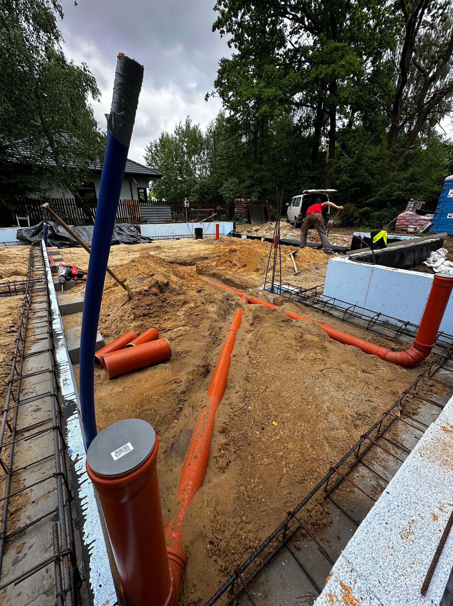 Instalacja rur kanalizacyjnych w wykopie fundamentowym, widoczne zbrojenie, izolacja termiczna i pracownik wyrównujący teren.