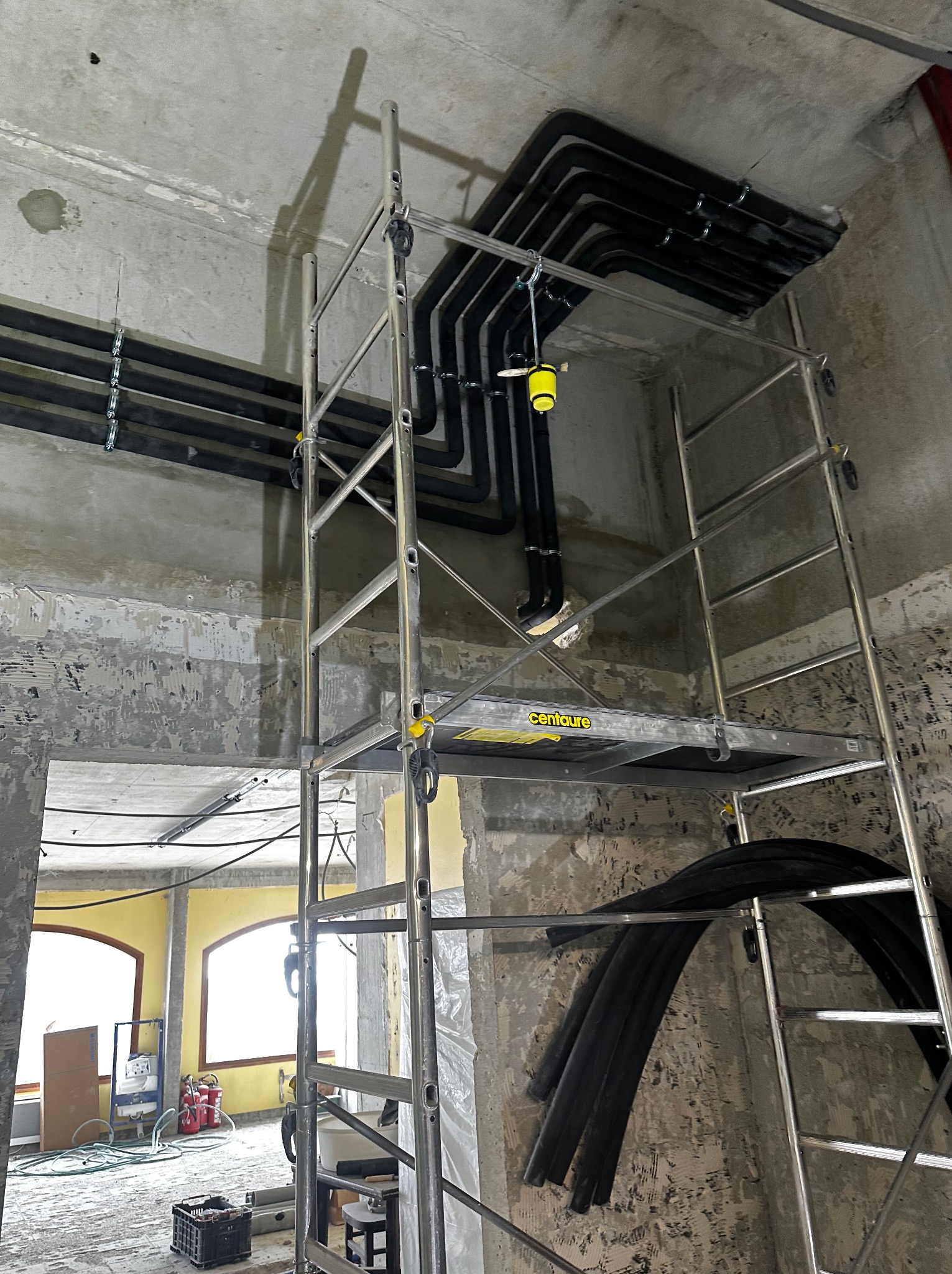 Montaż instalacji rurowej pod sufitem w surowym wnętrzu, widoczne rusztowanie i fragmenty nieotynkowanych ścian.