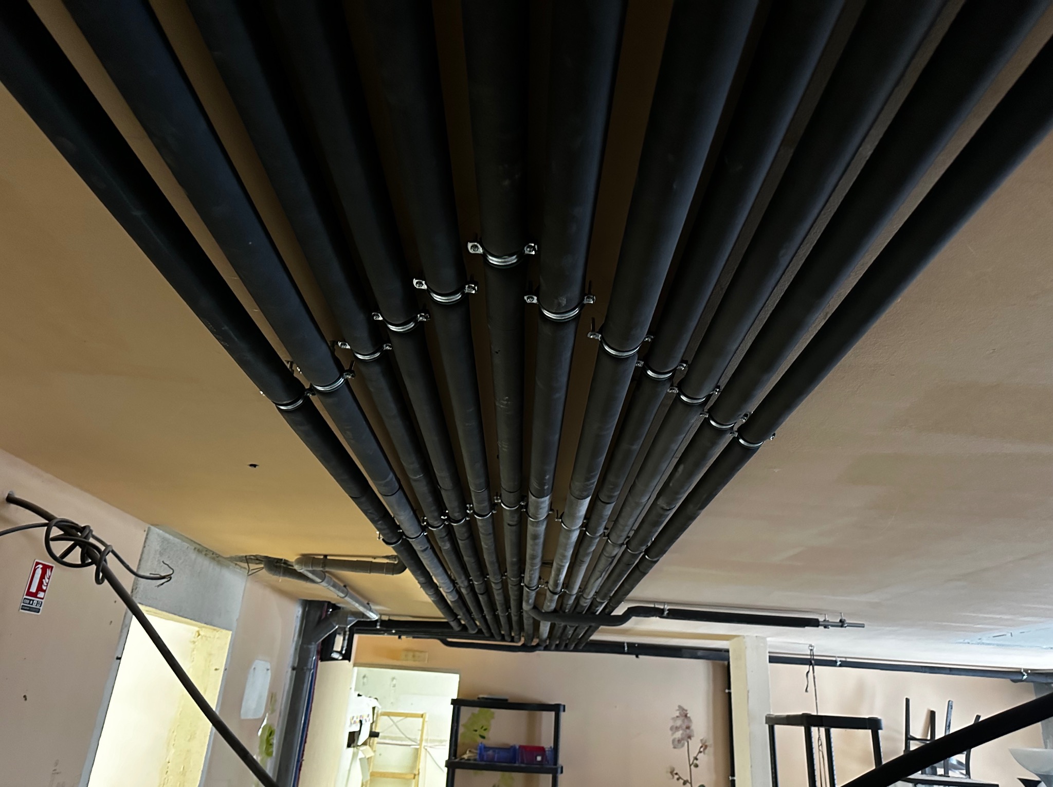 Równoległe czarne rury instalacji hydraulicznej pod sufitem, mocowane srebrnymi obejmami, widok z dołu, fragment pomieszczenia z gaśnicą w tle.